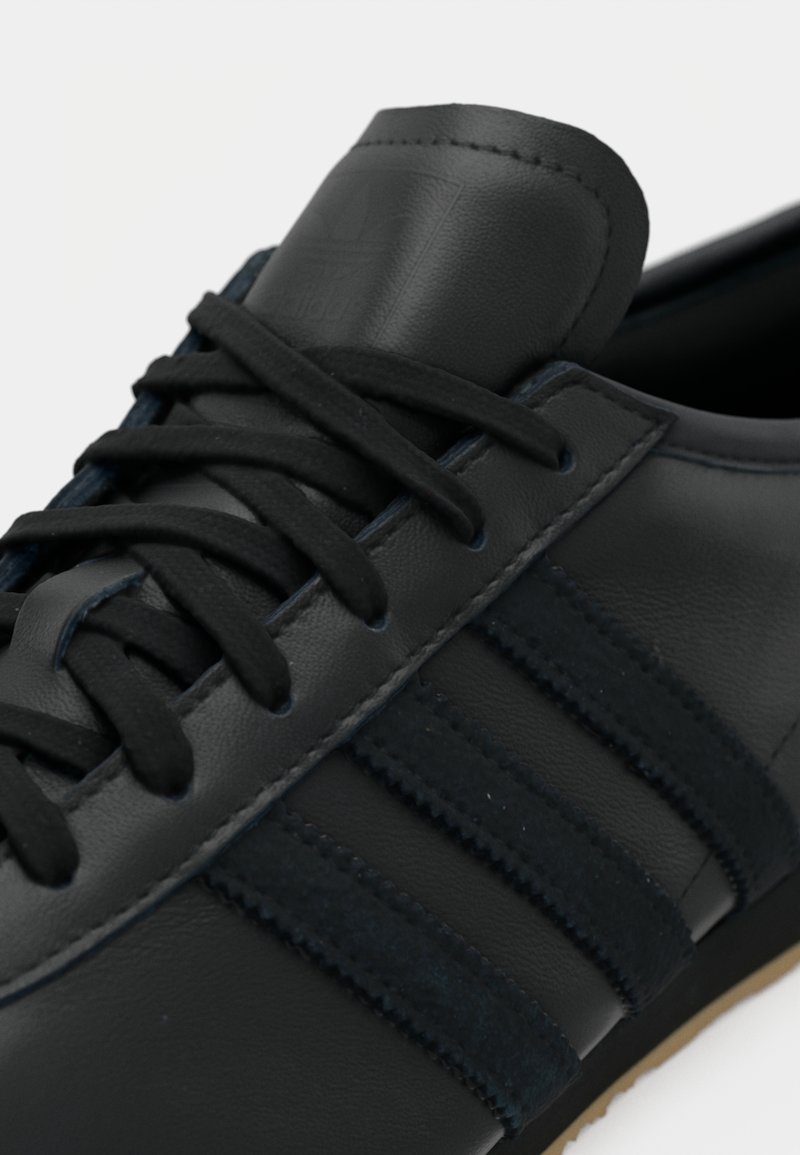 Black leather sneaker with dark navy suede stripes. Features black laces and a padded tongue with embossed logo. Gum rubber sole.