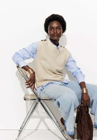 Gilet en tricot beige avec boutons d'épaule sur une chemise à rayures bleu clair. Jean bleu clair et sac en daim marron. Accents de bijoux en argent.
