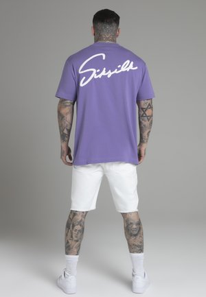 Purple cotton t-shirt with white script text "Sissilk" on the back, paired with white denim shorts and white sneakers. Visible tattoos on legs.