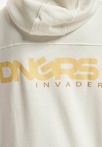 Fehér kapucnis pulóver, nagy arany grafikával, amely a hátán "DNGRS INVADER" feliratot olvasható. Puha textúrájú, laza szabású és kapucnis dizájnnal rendelkezik.