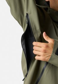Oliven grøn vandtæt jakke med en sidelukket lomme. Har glat stof, slank design og en tommelhul ved manchetten.