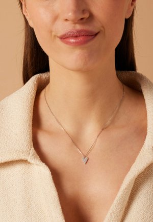Gros plan d'une femme portant un collier pendentif en forme de cœur en argent et une chemise beige à col texturé.