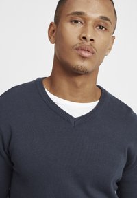 Maglione blu navy a scollo a V realizzato in tessuto a maglia testurizzato, indossato sopra una t-shirt bianca a girocollo. Design semplice e funzionale, senza loghi visibili.