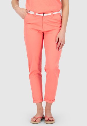 Frau trägt korallenrote Hose mit umgekrempeltem Saum, weißen gewebten Gürtel und korallenrote Sandalen, steht mit einer Hand in der Tasche vor einem schlichten Hintergrund.