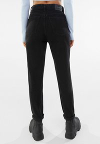 Person som bär högmidjade svarta jeans med uppvikta fållar, svarta klumpiga stövlar och en ljusblå långärmad topp, står mot en vit bakgrund.