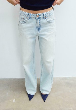 Oseba, oblečena v svetlo modre kavbojke z visokim pasom in ravnimi hlačnicami ter mornarsko modre pete s koničastim sprednjim delom, stoji ob navadni svetli steni.