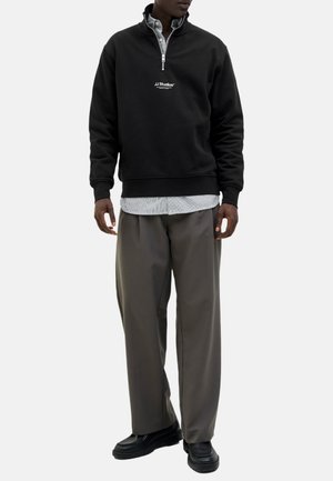 Schwarze Zip-up Sweatshirt mit gesticktem Logo, über einem gestreiften Hemd getragen. Lockere graue Hose und schwarze Schuhe ergänzen das Outfit.