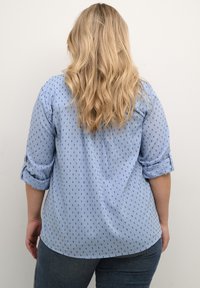 Camisa de botones de color azul claro con un patrón de puntos blancos y marinos, mangas enrolladas y dobladillo curvado. Tela suave con un corte relajado.