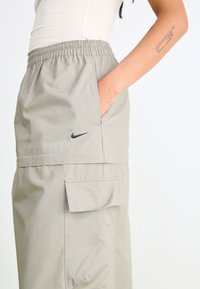 Svetlošedé cargo nohavice s elastickým pásom, bočnými vreckami a malým čiernym logom Nike na ľavom stehne. Hladká textúra tkaniny.