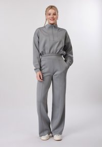 Jeune femme aux cheveux blonds portant un haut gris à manches longues avec fermeture éclair et un pantalon large assorti, debout avec une main dans la poche, souriante.