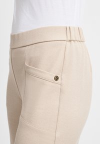 Beige Jogginghose mit weichem Material, elastischem Bund und dekorativer Seitentasche mit goldfarbenen Hardware-Details.
