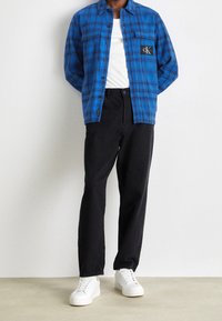 Blue plaid shirt with two chest pockets, black label detail; paired with black trousers and white sneakers. Smooth texture, relaxed fit.