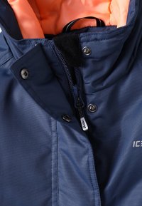 Marinblå vattentät jacka med en kontrasterande orange foder, försedd med dragkedja, tryckknappar och en mjuk krage med fleecekant.