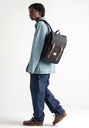Black backpack with brown leather accents, featuring a flap closure and metal hardware. Worn by a person in a light denim jacket and dark pants.