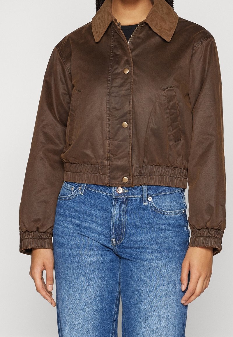 Veste courte marron avec poignets et taille élastiques, boutons-pression et poches avant inclinées, portée avec un jean bleu en denim.