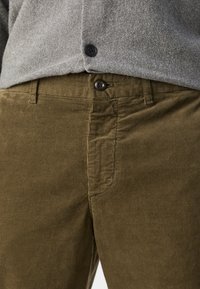 Pantalons en velours côtelé vert olive avec une texture côtelée, dotés de poches avant et d'une fermeture à bouton sombre. Portés sous un pull gris à boutons.