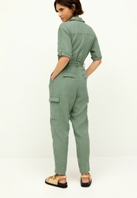 Zelený lněný overal s krátkými rukávy, páskem v pase a cargo kapsami. Sandály s texturovanými popruhy doplňují outfit.