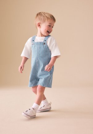Young boy in white shirt and blue striped overalls walking and smiling against a plain beige background