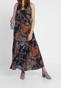 Kvinna i en ärmlös maxiklänning med ett flerfärgat blommigt paisleymönster, bärande på en lavendelfärgad axelväska och med matchande klacksandaler.