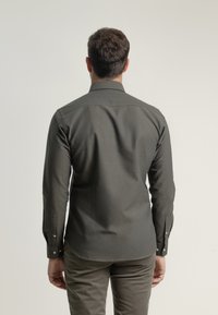 Camisa de manga larga en verde oscuro con cuello tipo camisa, puños abotonados y una tela suave y texturizada. La vista posterior muestra un dobladillo redondeado.
