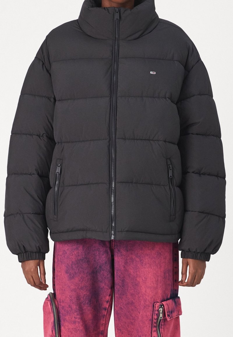 Veste matelassée noire avec col haut, fermeture éclair et design matelassé horizontal. Comprend deux poches latérales zippées.