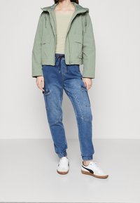 Person klädd i en ljusgrön huoodjacka, beige topp, blå denim cargo-joggers och vita sneakers med svart rand.