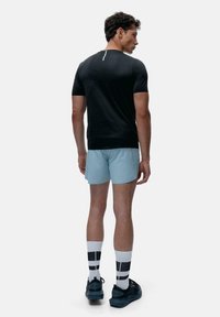 Melnā sporta t-kreklā, gaiši zilos šortos, baltās un melnās rakstainās zeķēs un melnās kurpēs. Modelis redzams no aizmugures pret vienkāršu fona.