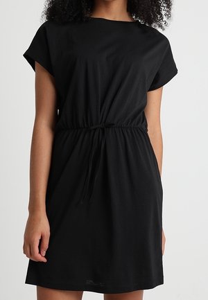 Kobieta w czarnej sukience z krótkim rękawem i paskiem podkreślającym talię, stojąca na jednolitym jasnym tle.