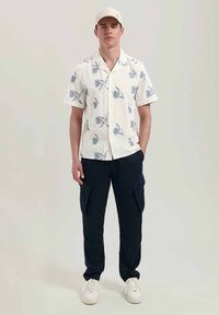 Camicetta bianca a maniche corte con motivo floreale blu; abbinata a pantaloni cargo blu scuro e sneakers bianche; un cappellino semplice completa il look.