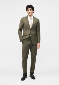 Άνδρας που φοράει ελιάς πράσινο κοστούμι με λευκό πουκάμισο, μπεζ γραβάτα και μαύρα παπούτσια, στέκεται μπροστά σε ένα λευκό φόντο.