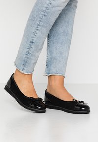 Bailarinas negras con punta brillante, cuerpo de ante y un lazo decorativo. Cuentan con una suela de goma plana y un diseño redondeado. Se llevan con jeans recortados.