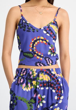 Femme portant un haut court sans manches bleu et un pantalon assorti avec des motifs abstraits multicolores en pointillés, les mains dans les poches.