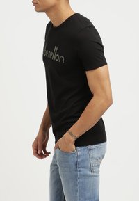 Černé bavlněné tričko s kulatým výstřihem a krátkými rukávy. Na přední straně má výrazné texturované logo. Je kombinováno se světle modrými džíny.