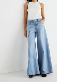 Jeans azzurri a gamba larga con vita alta, chiusura con bottone e tasche frontali, abbinati a una canotta bianca a coste e scarpe nere con borchie.