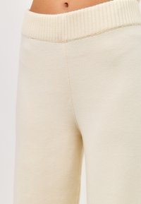Pantaloni de trening tricotati de culoare crem, cu un brâu lat cu pliuri și o textură moale, având un croi relaxat și fără buzunare sau embellishments vizibile.