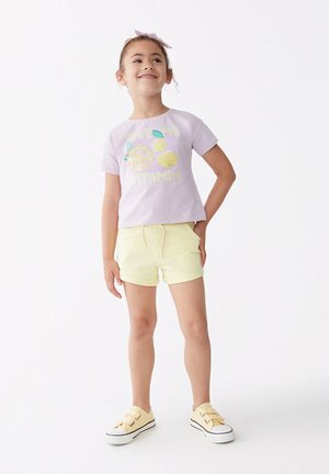 Camiseta de manga corta morada clara con gráficos de limones y texto amarillo, combinada con pantalones cortos amarillos. Zapatillas blancas con acentos negros.
