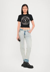 Kratinta juoda marškinėliai su logo, šviesiai mėlynos nusidėvėjusios džinsai ir juodi kariniai batai. Modelis laiko mažą juodą rankinę.