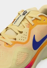 Chaussure de course Nike jaune et orange avec des lacets beige, un logo swoosh bleu et une étiquette "RUN structure 26" sur la languette.