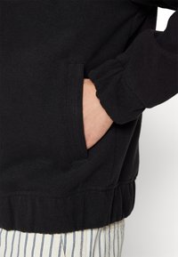 Sort sweatshirt med tekstureret stof, elastisk talje og sidelomme. Ærmerne er samlet, hvilket giver en tætsiddende pasform.