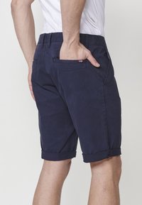 Man in een donkerblauwe opgerolde shorts en een wit shirt, met de hand in zijn achterzak, wat een casual zomerse outfit laat zien.