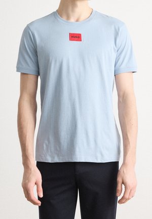 Mężczyzna ubrany w jasnoniebieski T-shirt z krótkim rękawem, z czerwonym logo „HUGO” na piersi, oraz czarne spodnie, stoi na jednolitym tle.