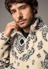 Fleece pullover i creme med sorte blomster- og geometriske mønstre, som har en halv lynlås krave og tekstilt, blødt stof.