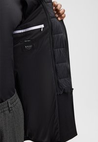 Mano che tiene aperto un cappotto di lana nera, rivelando una fodera trapuntata con tasca bianca con zip e etichette di tessuto all'interno.