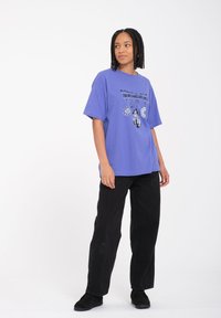 Camiseta oversized morada con estampado gráfico de motivos celestiales, combinada con jeans negros de pierna recta y zapatos negros.
