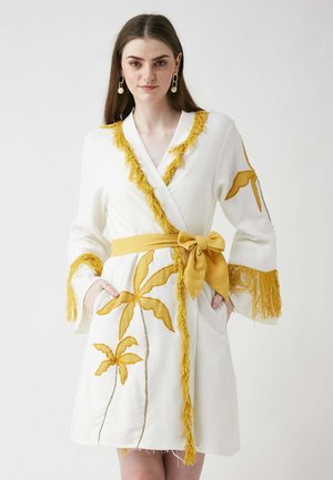 Weißes Gewand mit gelben Fransen, floraler Stickerei und passendem Gürtel. Verfügt über lange Ärmel und seitliche Taschen. Strukturierte Details heben das Design hervor.