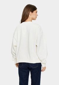 Fehér, oversize pulóver leengedett vállakkal és bordás szegéllyel, sötétkék farmernadrággal párosítva. Sima textúra, minimális dizájn.