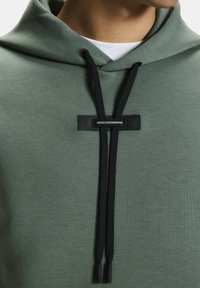 Zöld kapucnis pulóver texturált felülettel, fekete húzózsinórral és a középen elhelyezkedő fekete címkével, amelyen az olvasható: "SWISS ENGINEERING".