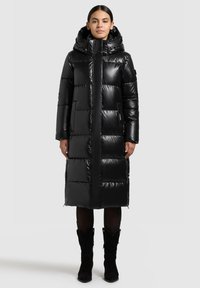 Duga crna jakna na punjenje sa sjajnim završetkom, velikim kapuljačom, patentnim zatvaračem i vodoravnim prošivnim uzorkom. Ima bočne džepove i duljinu do sredine bedara.