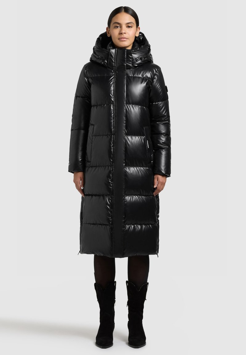 Duga crna jakna na punjenje sa sjajnim završetkom, velikim kapuljačom, patentnim zatvaračem i vodoravnim prošivnim uzorkom. Ima bočne džepove i duljinu do sredine bedara.