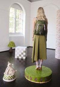 Olivovo zelený batoh s levanduľovými akcentmi, nosený cez dlhé olivovo zelené šaty. Žlté lodičky a biele členkové ponožky. Dekorácia zahŕňa rozprávkové sochy húb a zajacov.